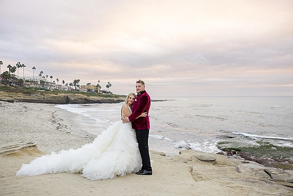 Couple portrait at a beach wedding portraits session, bride with long train leaning on groom in velvet jacket by rocky cliffs and ocean