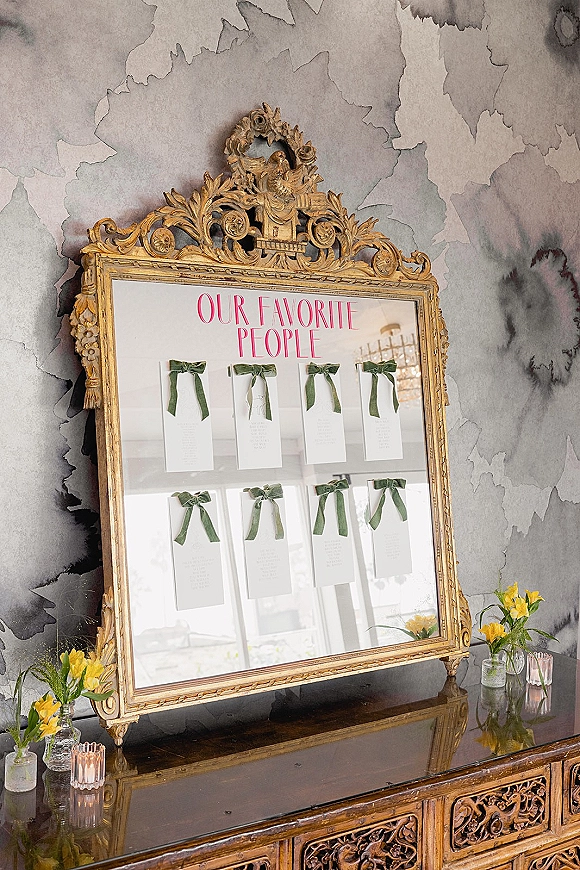 Escort card display on an ornate gold mirror with green ribbon bows, yellow bud vases, and votive candles on a carved console table.