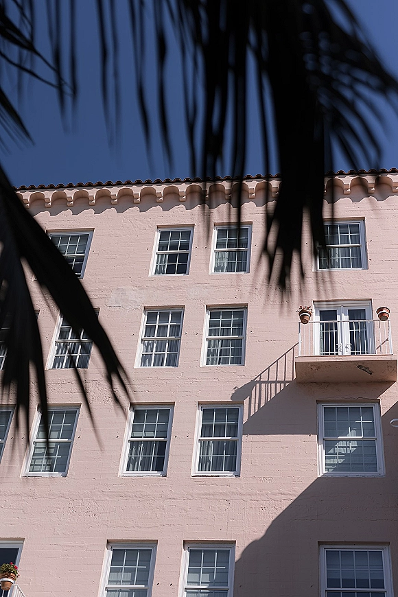Wedding venue exterior with a small balcony and potted plants on a pale blush stucco facade under blue sky and sharp sunlit shadows