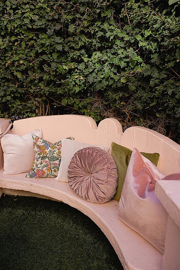 Wedding lounge seating with a curved upholstered sofa and mix of velvet and floral pillows set on grass against an ivy hedge wall