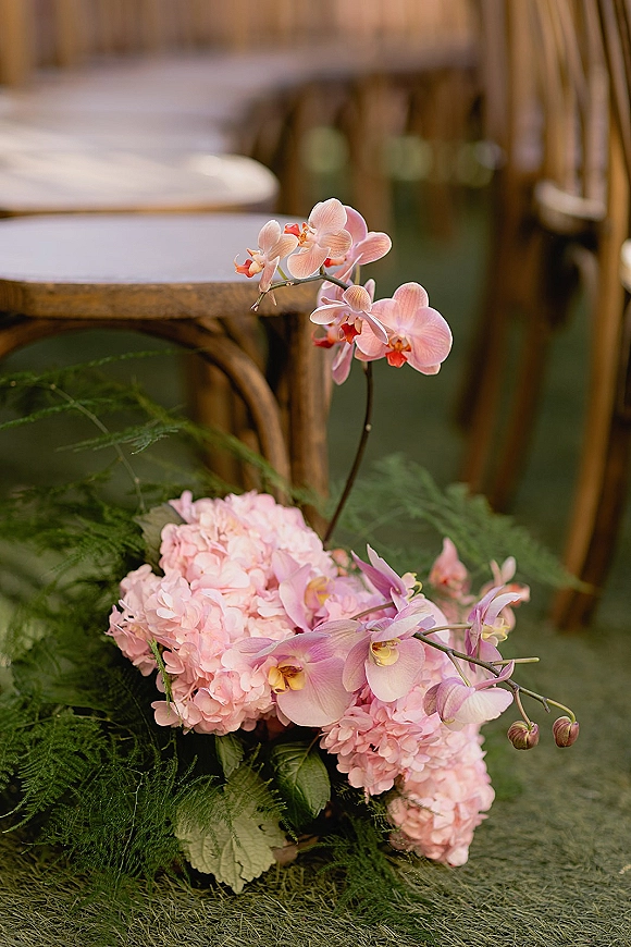 Wedding centerpiece of pink orchids and hydrangea with greenery on a wooden table, with reception seating softly blurred behind