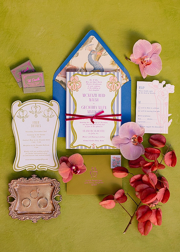 Invitation suite wedding invitation flat lay with blue envelope, striped cards, pink ribbon, orchids, rings on ornate silver tray over green texture