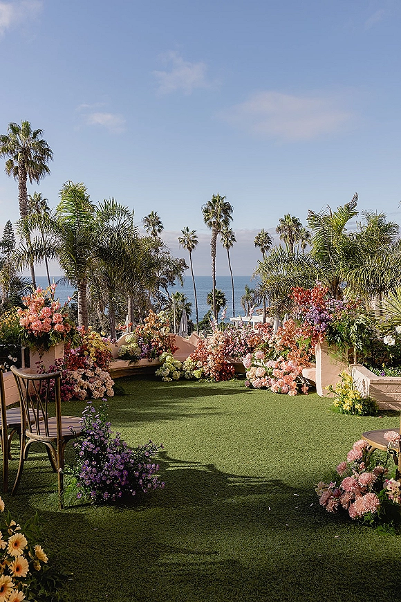 Outdoor ceremony setup with floral arrangements lining a green lawn runner and chairs, on a terrace with palm trees and ocean view