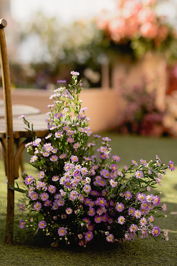Ceremony aisle florals with aisle marker flowers of purple daisies and greenery beside a rustic wooden chair on a grassy lawn
