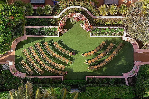 Ceremony setup for an outdoor wedding ceremony with semi-circle chairs around a floral arch on a garden lawn, framed by palm trees