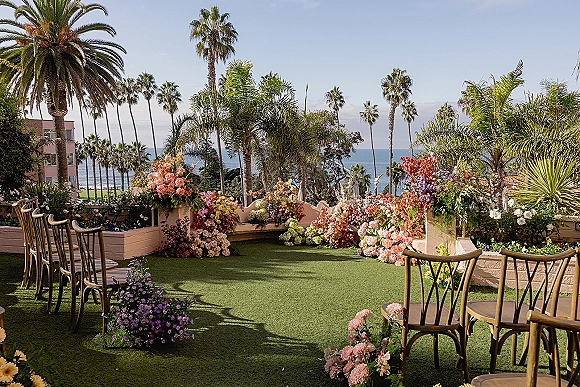 Ceremony setup for an outdoor wedding ceremony with pastel floral pillars and aisle blooms on a terrace overlooking palm trees and ocean view