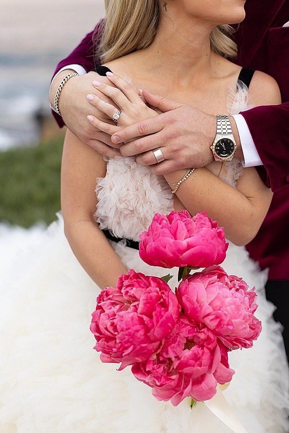 Engagement ring photo of a hand with engagement ring and wedding band, pale pink manicure, holding pink peony bouquet by water outdoors