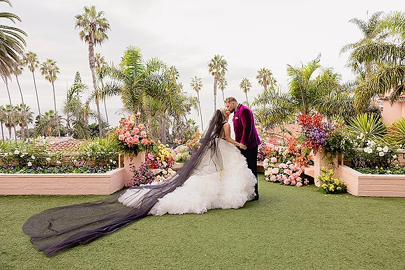 Wedding kiss portrait of bride and groom kissing, her black cathedral veil and long train flowing on a palm-lined garden terrace