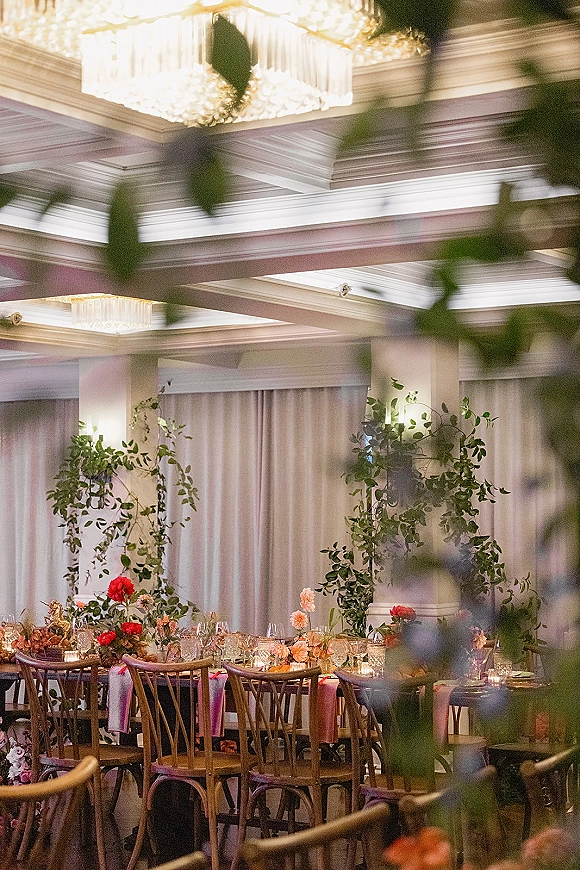Reception tablescape with wedding head table decor, farmhouse tables, floral centerpieces, greenery vines, taper candles, and chandelier-lit draped backdrop