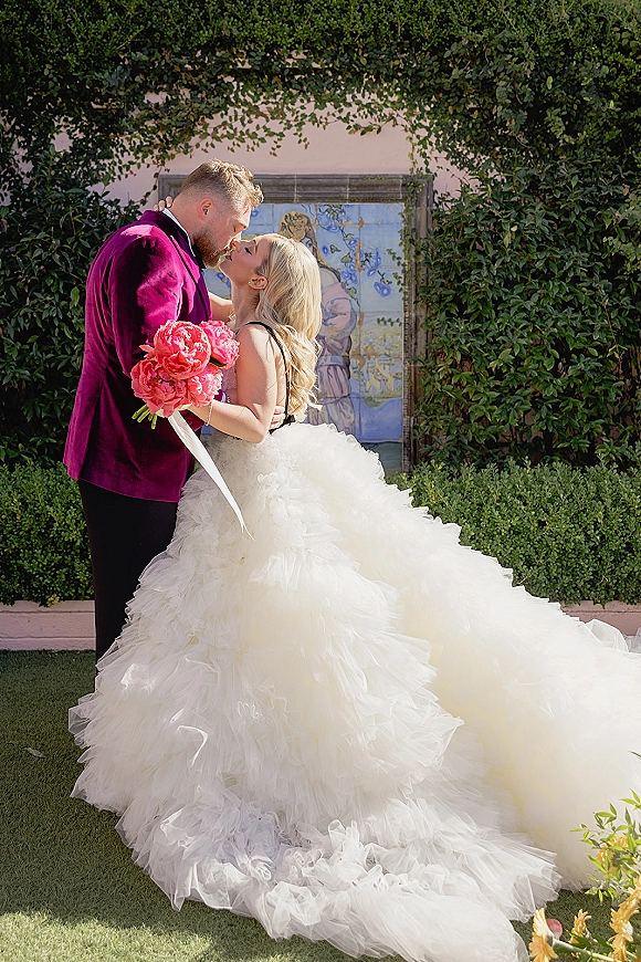 Wedding couple portrait of bride and groom kissing, bride holding a pink peony bouquet as her ruffle tulle train flows by an ivy wall