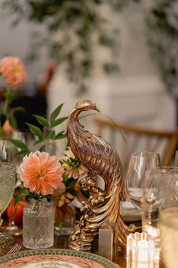 Reception tablescape with a gold bird centerpiece, peach flowers, ribbed glass votives, patterned plates, and gold flatware with white chairs behind