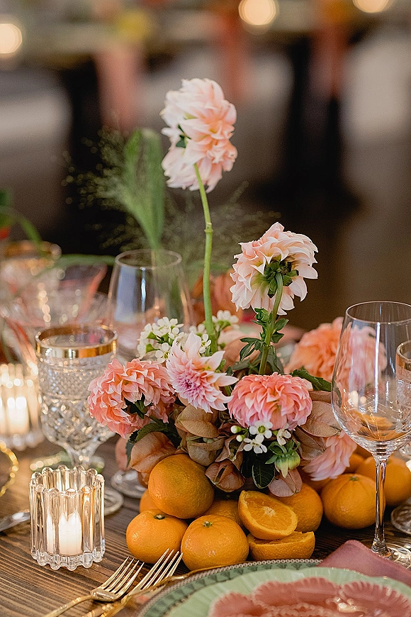 Reception tablescape with a wedding table centerpiece of pink dahlias and oranges, ribbed glass votives, and crystal glassware in warm light