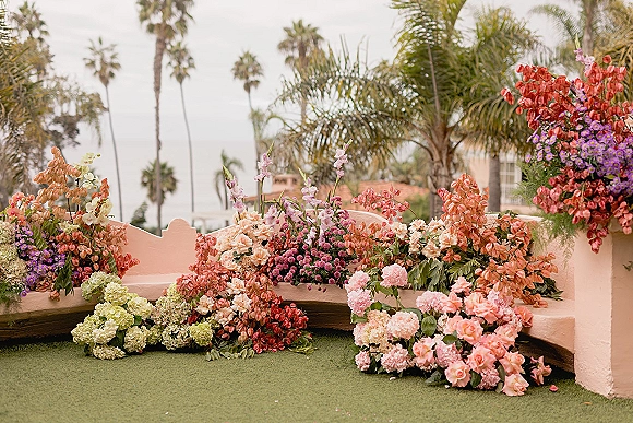 Ceremony floral decor with hydrangeas, roses, orchids and hanging amaranthus, framing an ocean-view lawn with palm trees behind