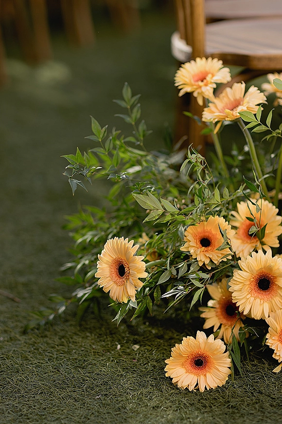 Aisle florals with ceremony aisle flowers in yellow gerbera daisies and greenery beside a wooden chair on a grass lawn