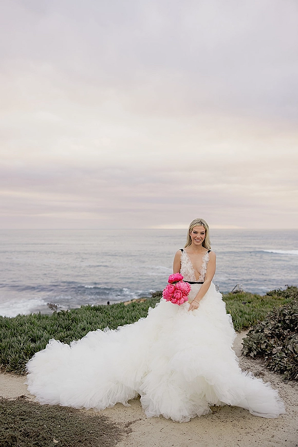 Bridal portrait of a bride in a tulle ball gown with black straps and waistband, holding a pink peony bouquet on ocean cliffs under clouds