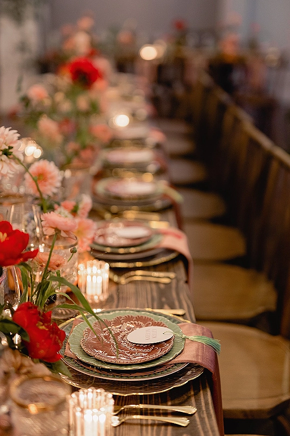 Reception tablescape with wedding table place setting, layered plates, tassel place cards, votive candles, and floral centerpieces on a long banquet table