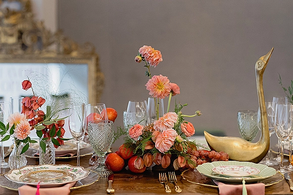 Reception tablescape with wedding table centerpiece of peach flowers, greenery, bud vases and grapes, set with gold flatware and vintage china under an ornate mirror