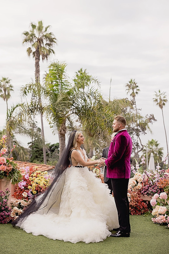 Wedding vows as bride and groom hold hands outdoors, her long black veil flowing beside palm trees and colorful floral arrangements