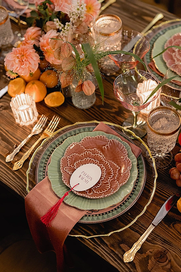 Reception tablescape with layered dinner plates, gold flatware, patterned glassware, votive candles, and floral centerpiece on a wood table