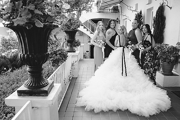 Bridal party portrait of a bride with bridesmaids in black dresses holding bouquets on an outdoor veranda by white stairs and planters