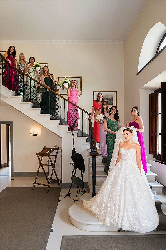 Bridal party portrait of bride in lace gown and veil with bridesmaids holding bouquets and champagne on an indoor staircase