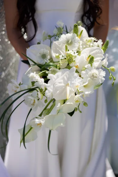 Bridal bouquet with white orchid bouquet blooms and calla lilies, cascading with green foliage and ribbon against outdoor light and blurred wedding party