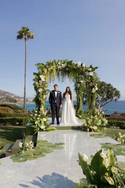 Ceremony setup with an outdoor wedding altar, floral arch and white aisle runner with lily pads beside an oceanfront lawn under blue sky