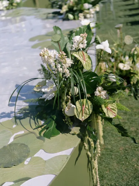 Reception tablescape with a green and white tablescape runner of lush greenery and white flowers on a patterned cloth, set on an outdoor lawn