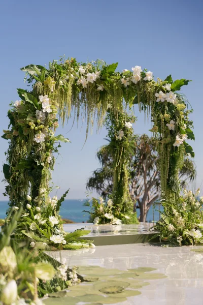 Wedding ceremony arch with white flowers and hanging greenery, set on a clear platform overlooking the ocean under blue sky
