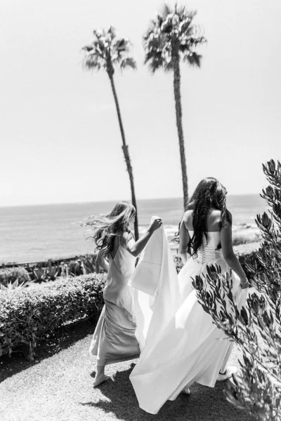 Bridal party moment as bride walks away while bridesmaid holds her veil, wedding dress train flowing on a coastal bluff with ocean and palms