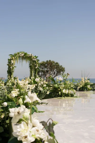 Ceremony aisle decor with glossy white runner leading to a floral arch of white blooms and greenery, set on an ocean-view lawn under blue sky