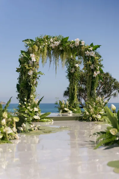 Wedding ceremony arch with a greenery wedding arch of white flowers and hanging greenery on a reflective aisle overlooking the ocean