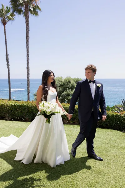 Couple portrait of bride and groom walking hand in hand, bride holding white bouquet, with ocean, palm trees, and blue sky behind