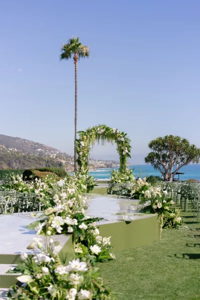 Ceremony setup with white floral arch and greenery garlands, lined with chairs and aisle florals on a lawn overlooking ocean coastline