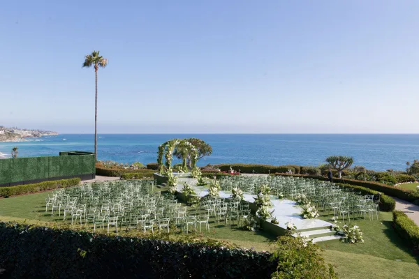 Ceremony setup for an oceanfront wedding ceremony with a white floral arch, clear platform stage, and flower-lined aisle on a lawn by the coast
