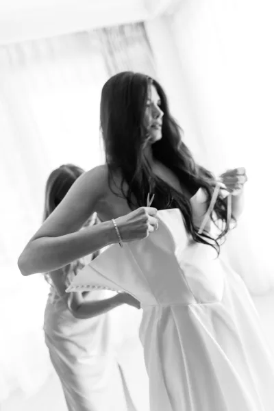 Bridal getting ready as bride putting on dress in a strapless gown, long wavy hair and bracelet, lit by a bright window and curtains