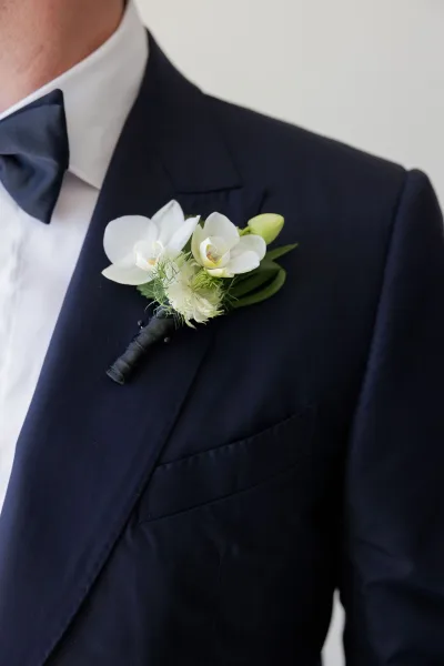 Wedding boutonniere featuring a white orchid boutonniere with greenery and black ribbon wrap on a tuxedo lapel against a neutral wall