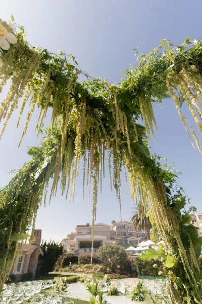 Wedding ceremony arch with greenery garland and hanging white florals, set on an outdoor lawn aisle with chairs under blue sky