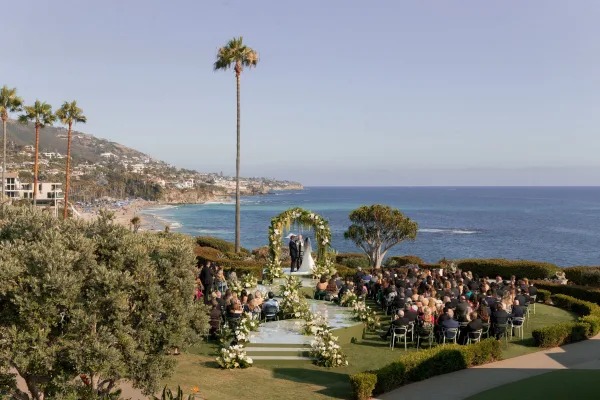 Outdoor wedding ceremony with a floral arch of white flowers and greenery, aisle florals and runner, set above the ocean cliffs with palms