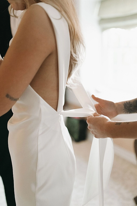Bridal dress detail of an open back wedding dress with bow straps on a white gown, softly lit by window light in an indoor room