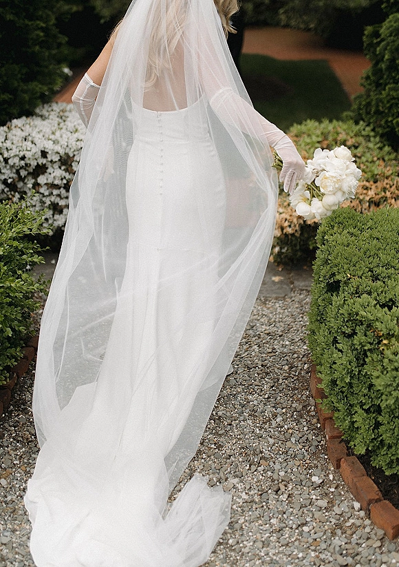 Bridal gown with cathedral veil flowing behind, strapless button-back dress and white peony bouquet on a garden gravel path