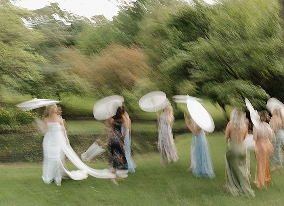 Bridal party photo of bride with bridesmaids holding white parasols, bouquet and long veil flowing on a green lawn by water