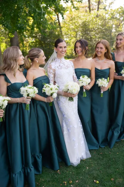 Bride with bridesmaids in emerald green bridesmaid dresses, holding matching bouquets as they stand together.