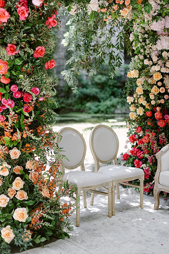 Wedding ceremony backdrop with a floral ceremony arch of coral and blush roses and hanging greenery, set in a garden along a stone walkway