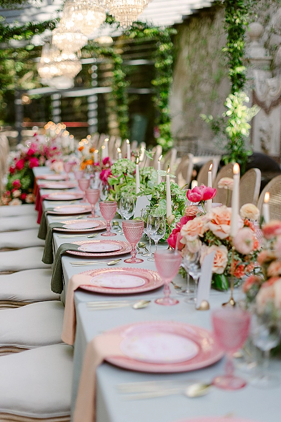 Reception tablescape with a long wedding reception table set with taper candles, garden rose florals, pink goblets, and chandeliers on an outdoor patio