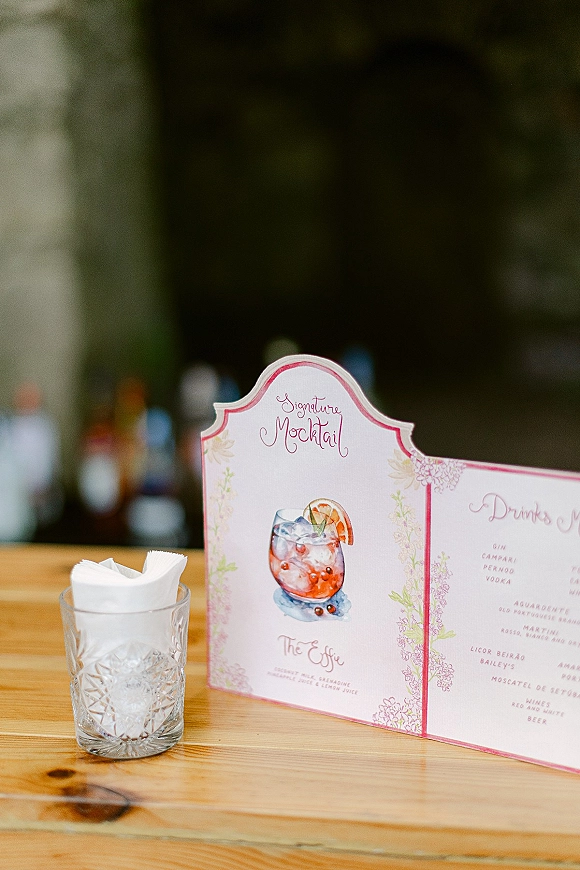 Wedding bar menu with signature cocktail sign on a wooden bar top, featuring watercolor drink art, pink border, and blurred bottles greenery behind