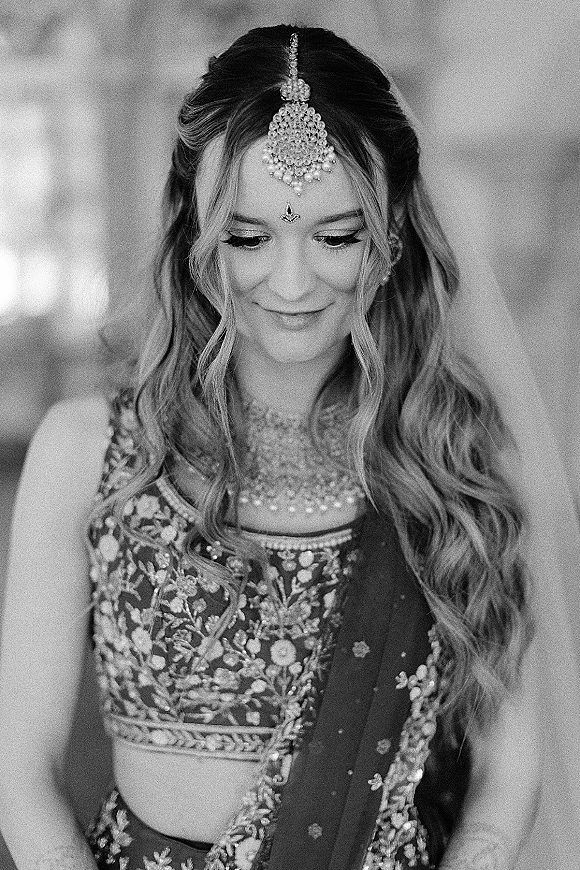 Bridal portrait of a South Asian bride wearing a statement maang tikka and ornate jewelry, with veil and soft glam makeup in blurred background