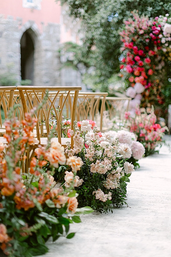 Ceremony aisle decor with wedding aisle flowers lining a stone walkway, wood chairs and rose-hydrangea clusters in a stone courtyard archway