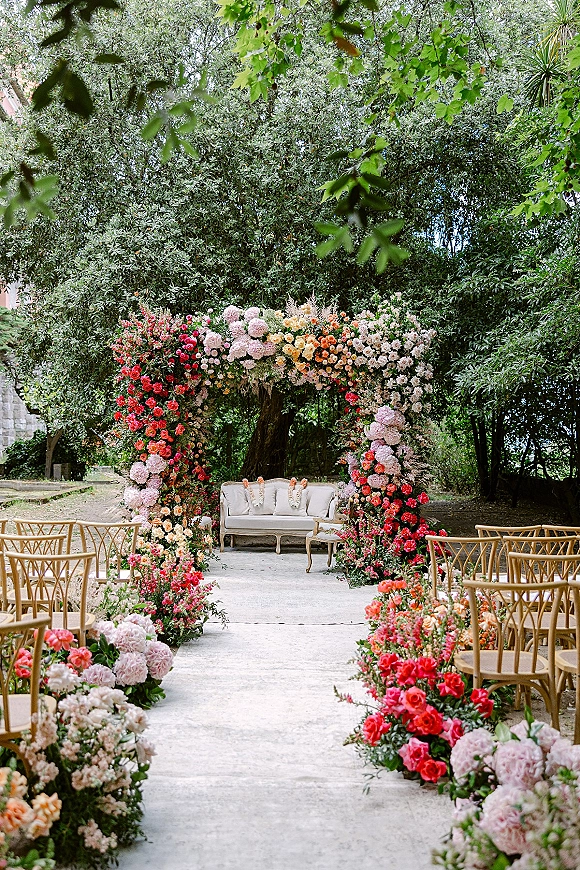 Ceremony setup with floral arch accents framing a white settee, rose and peony aisle flowers, and wooden chairs in a garden by a stone building