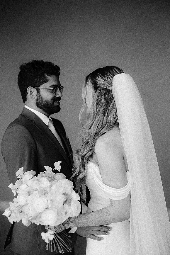 Couple portrait in a black and white wedding portrait, bride in off-the-shoulder dress with veil holding bouquet as groom in glasses faces her by a plain wall